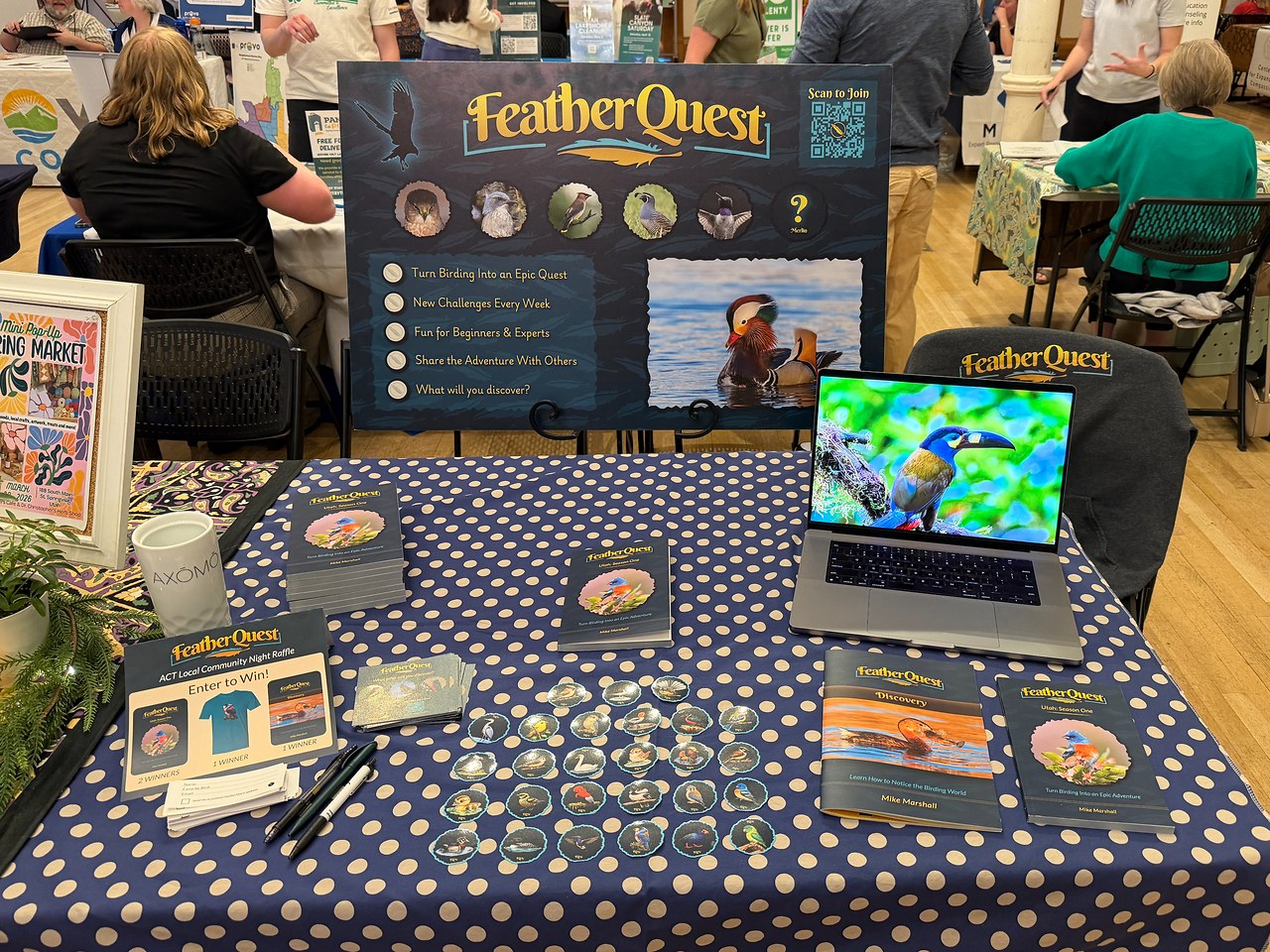 FeatherQuest booth at ACT Local Community Night showing the poster, stickers, books, and raffle table
