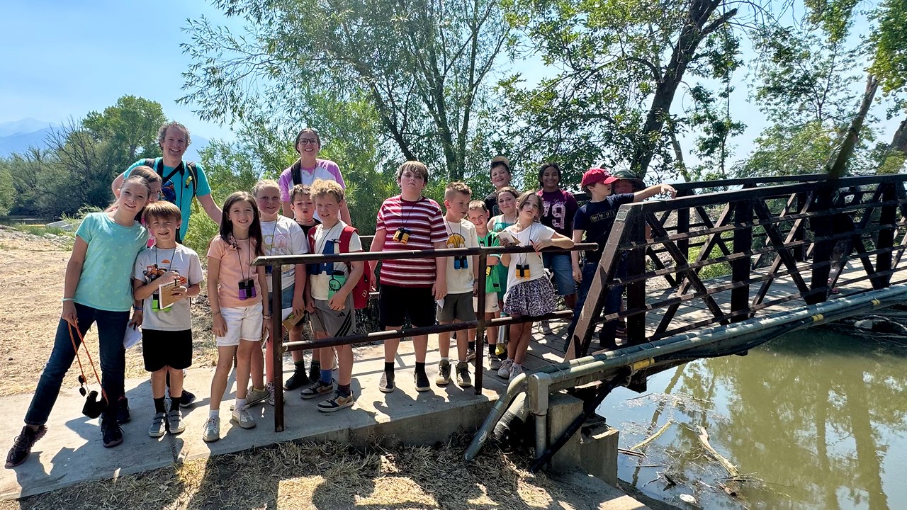 Nature school campers with binoculars gathered on a bridge during a FeatherQuest bird walk at Utah Lake State Park