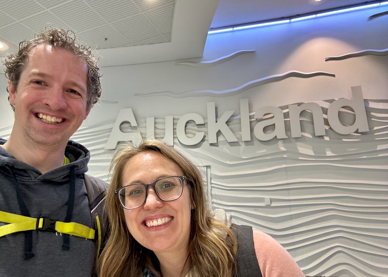Mike and his wife standing by an Auckland airport sign after arriving in New Zealand