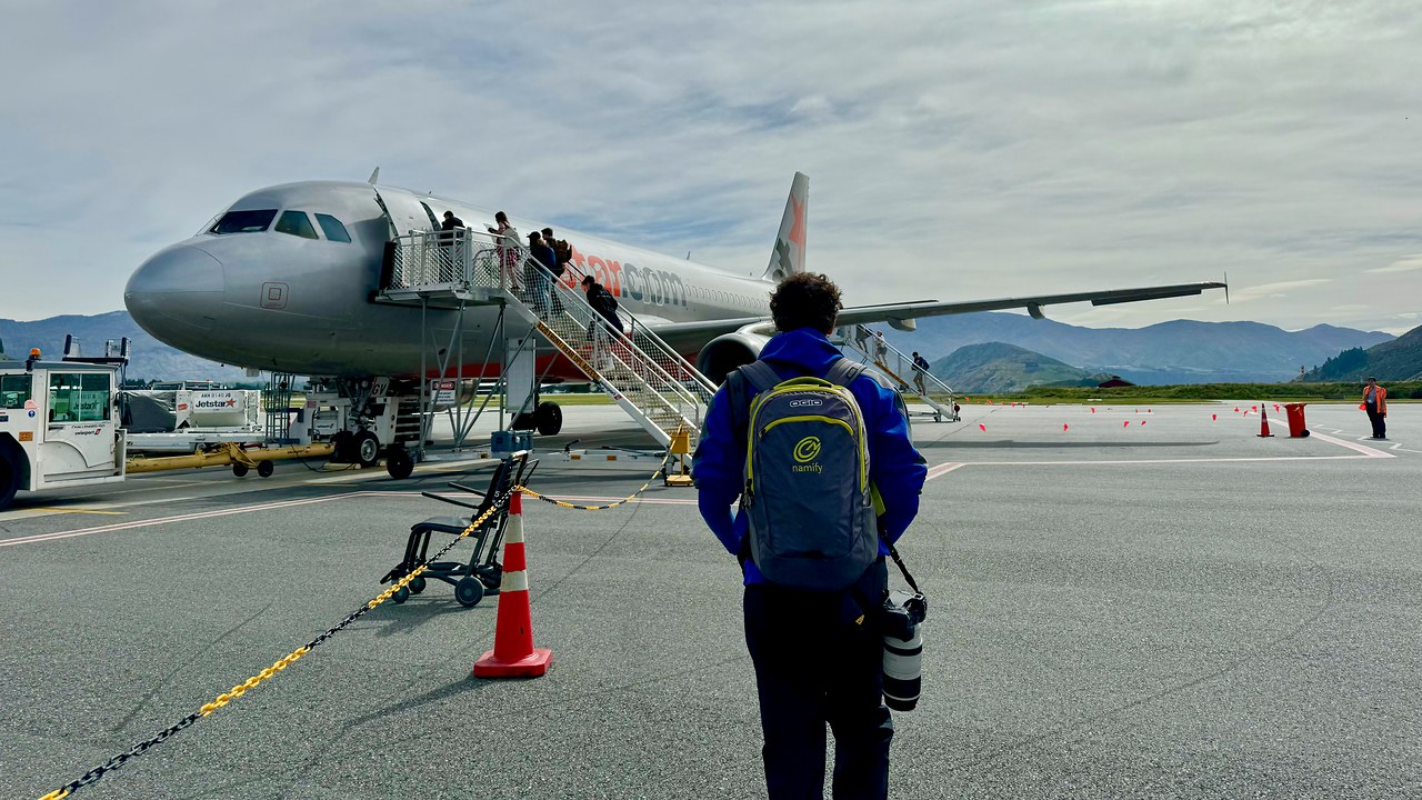 Walking toward an Air New Zealand plane with camera gear and mountains in the distance