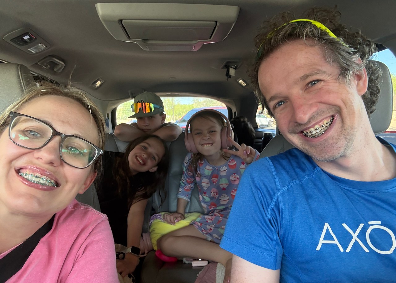 Mike and Stacy smiling for a road-trip selfie with their kids packed into the vehicle behind them on the way to Puerto Peñasco