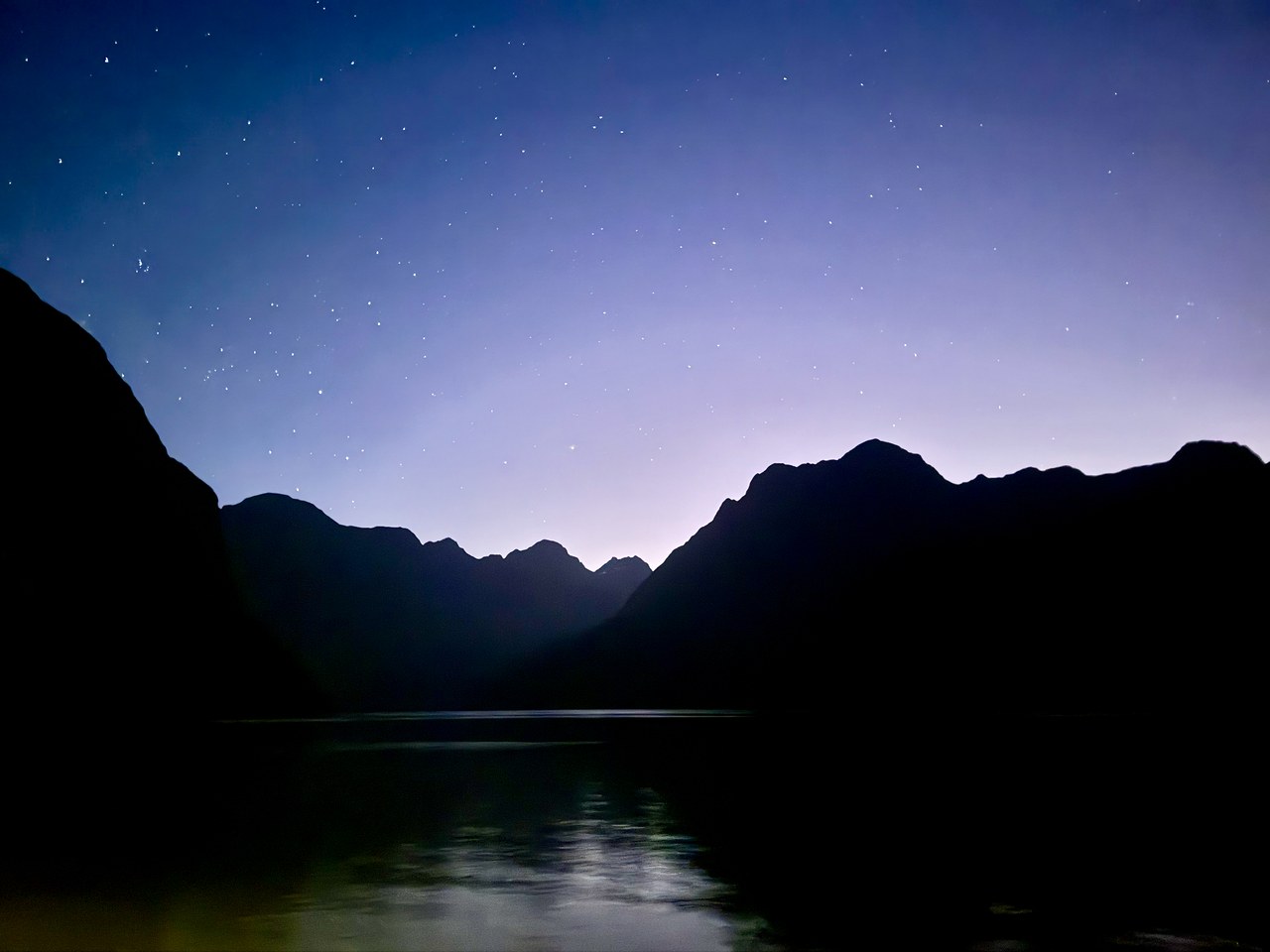 Stars shining above the silhouetted mountain walls of Milford Sound at night