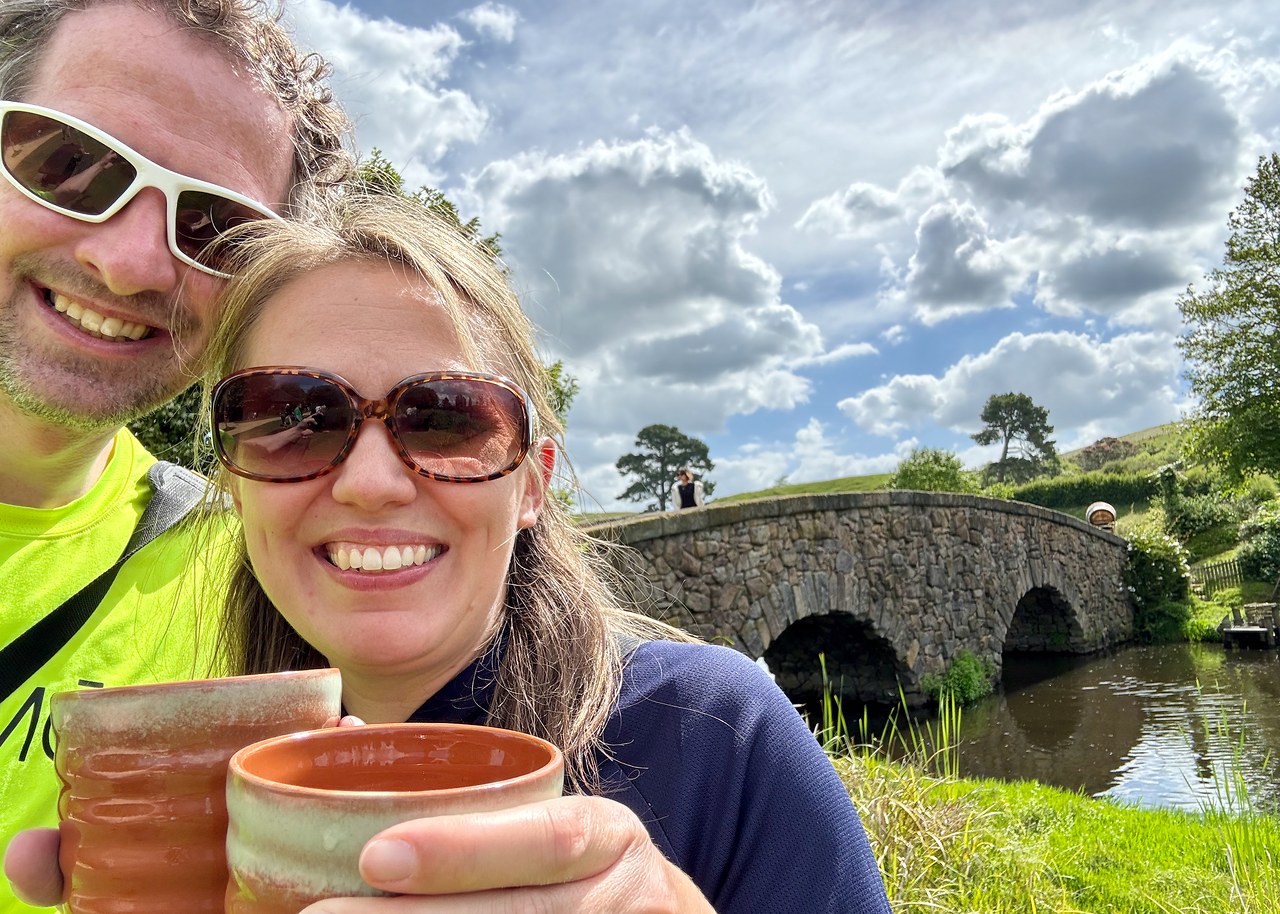Mike and Stacy holding mugs of non-alcoholic ginger beer beside the stone bridge at Hobbiton.