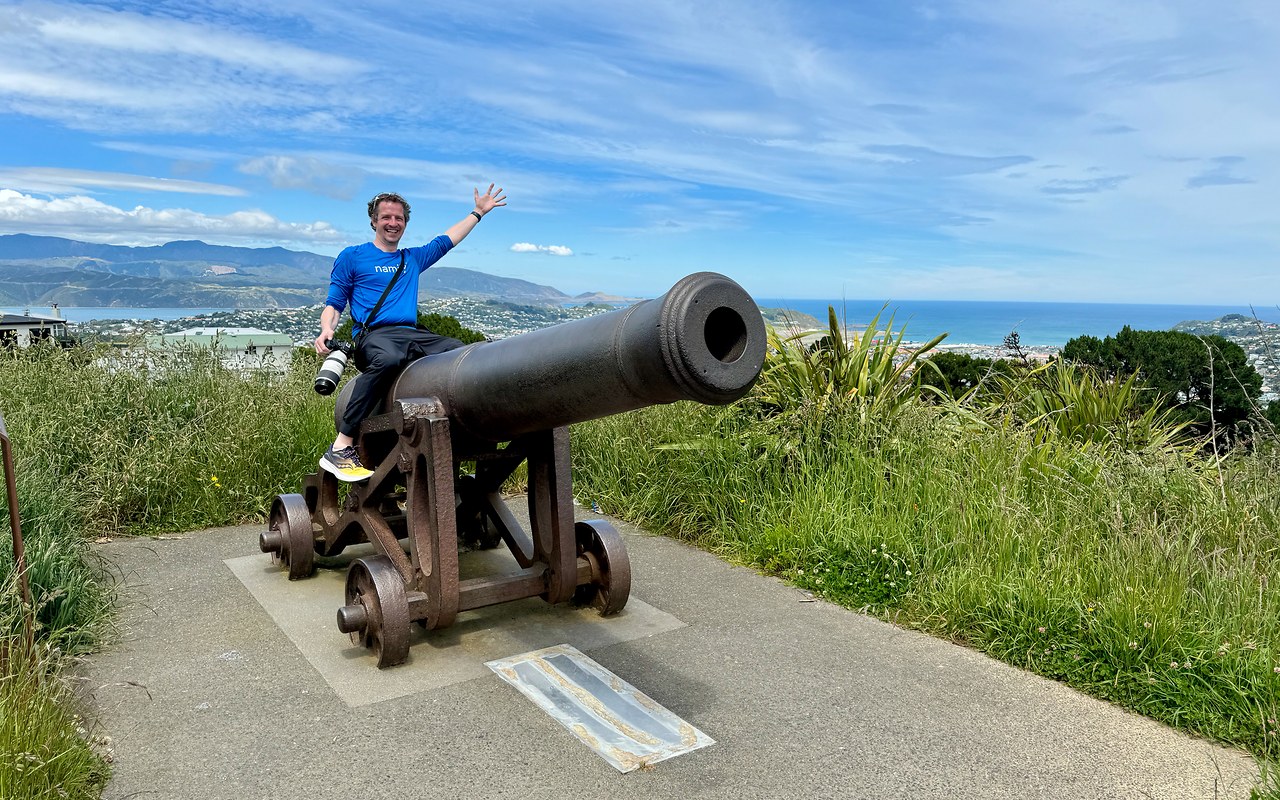 Mike sitting on a historic cannon at Mount Victoria Lookout with Wellington harbor and coastline behind him.