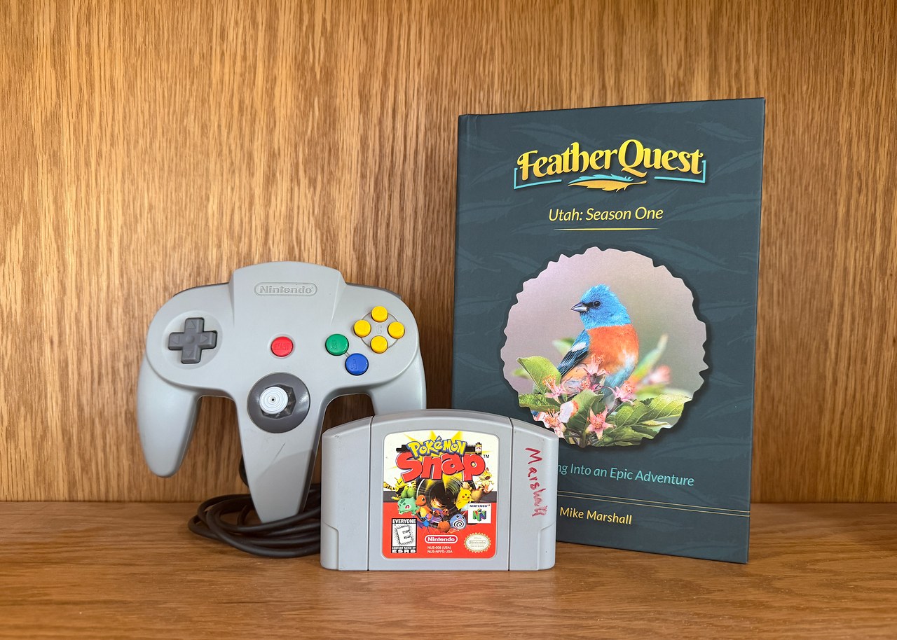 An original Pokémon Snap cartridge, Nintendo 64 controller, and FeatherQuest Utah: Season One book displayed together, representing the connection between childhood video games and real-world birding adventures.